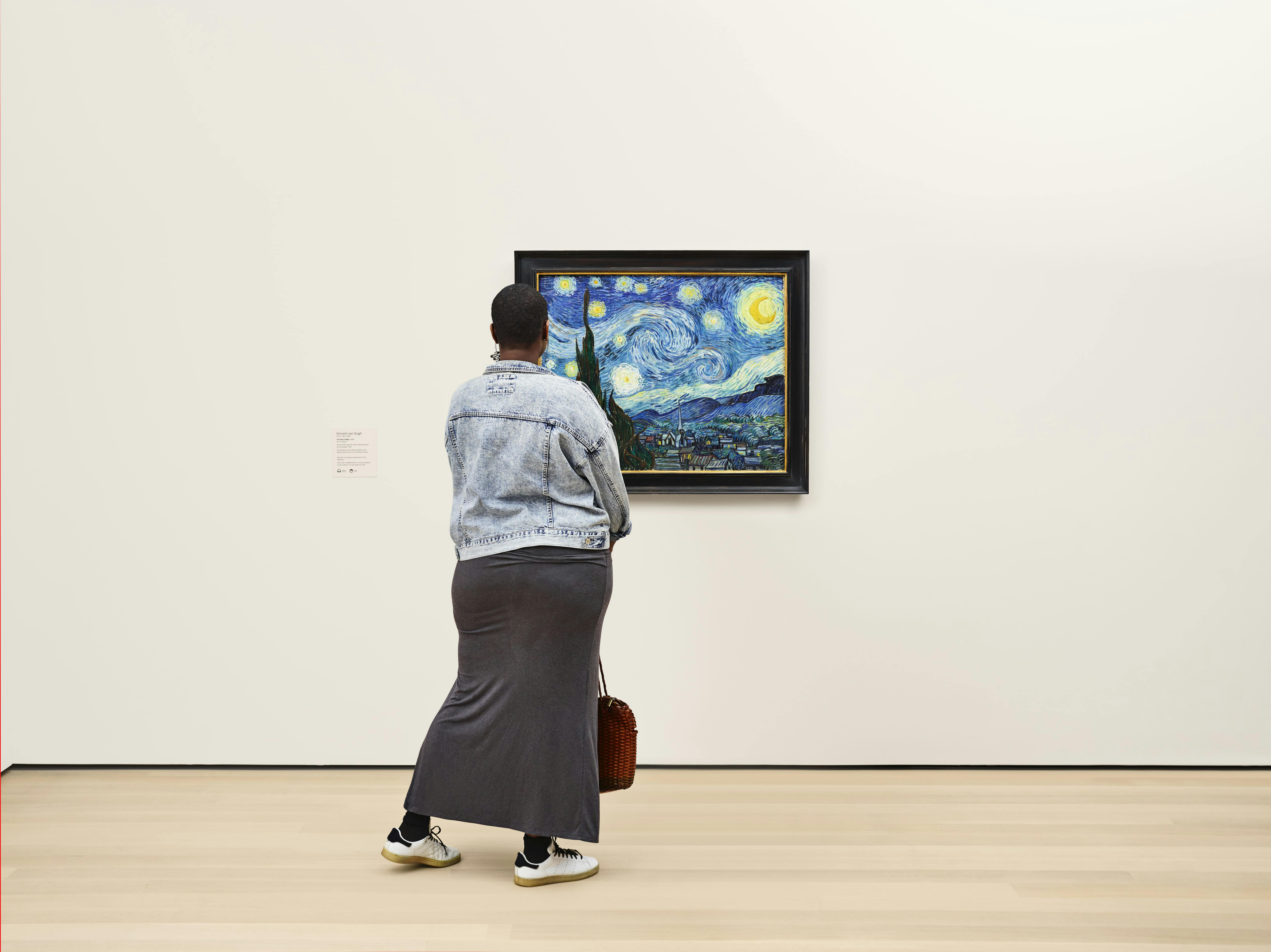 Uma pessoa com um casaco de ganga e uma saia comprida está em frente ao quadro "Noite Estrelada" de Van Gogh num museu.