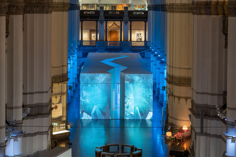 Large indoor space with tall columns, blue lighting, and a central white structure featuring abstract blue designs.