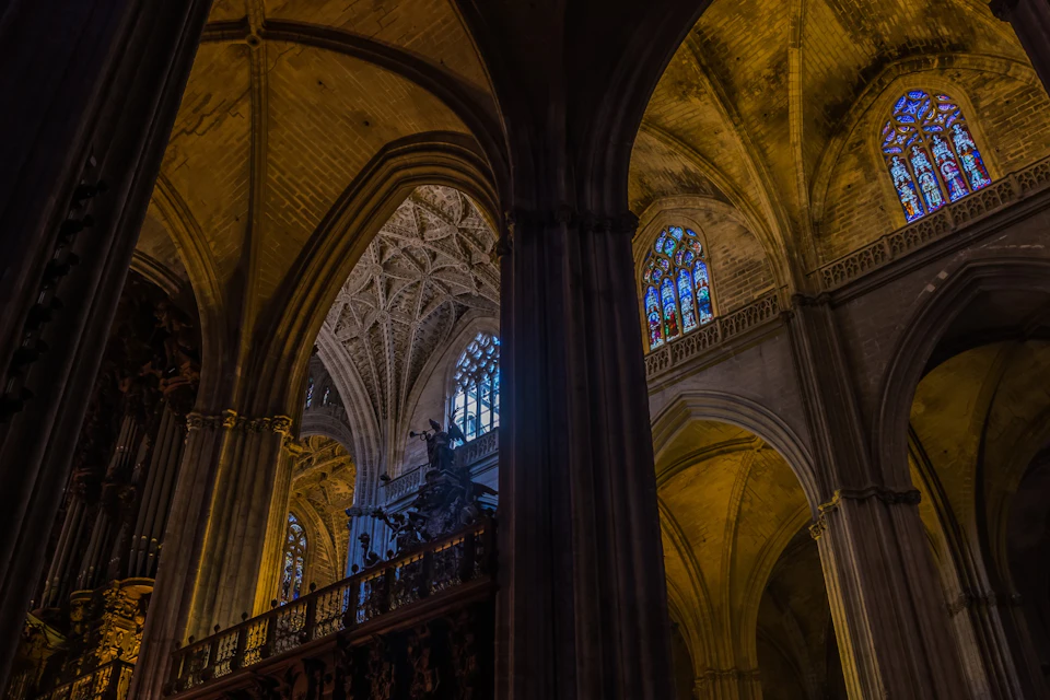 Interior of a cathedral with tall pillars, vaulted ceilings, and stained-glass windows illuminated by soft light.