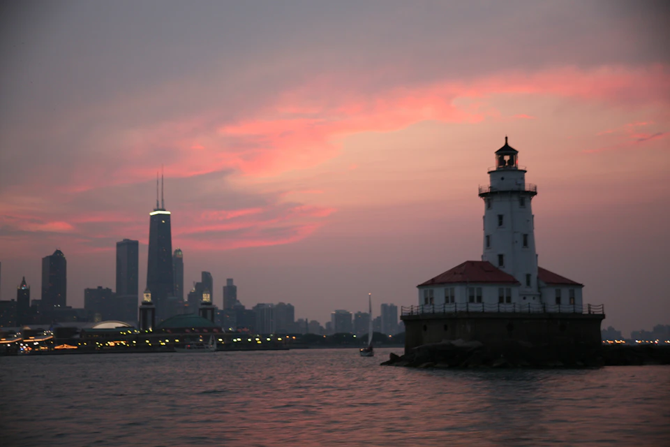 A lighthouse on the water at sunset with a city skyline and a tall, illuminated building in the background.