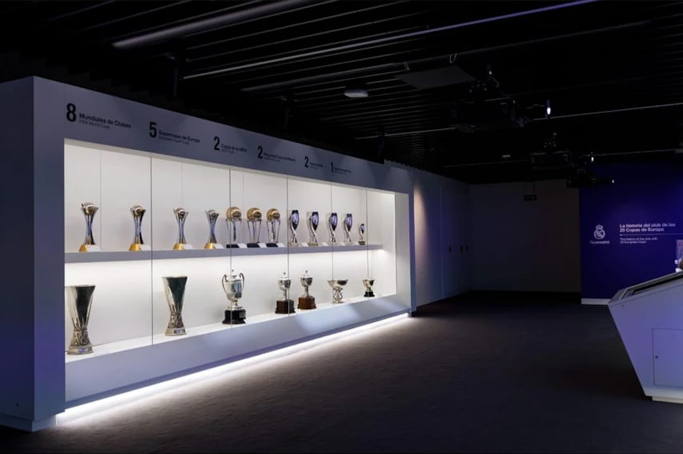 A dimly lit trophy room displaying various trophies on illuminated shelves, with labels above listing achievements.