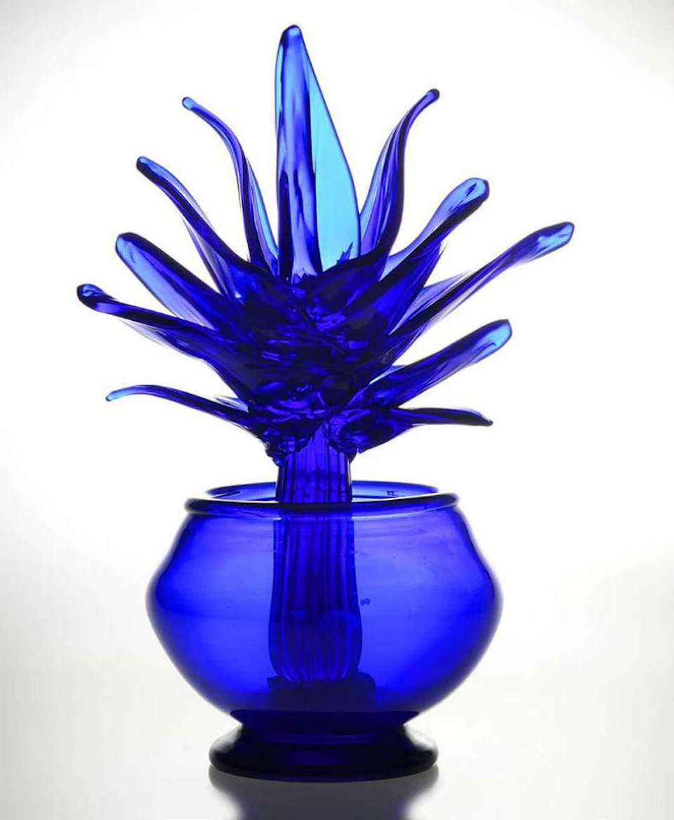 A bright blue glass sculpture resembling a spiky plant in a round vase, set against a plain white background.