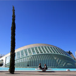 Två personer sitter på en bänk framför en modern, kupolformad byggnad med höga träd under en klarblå himmel.