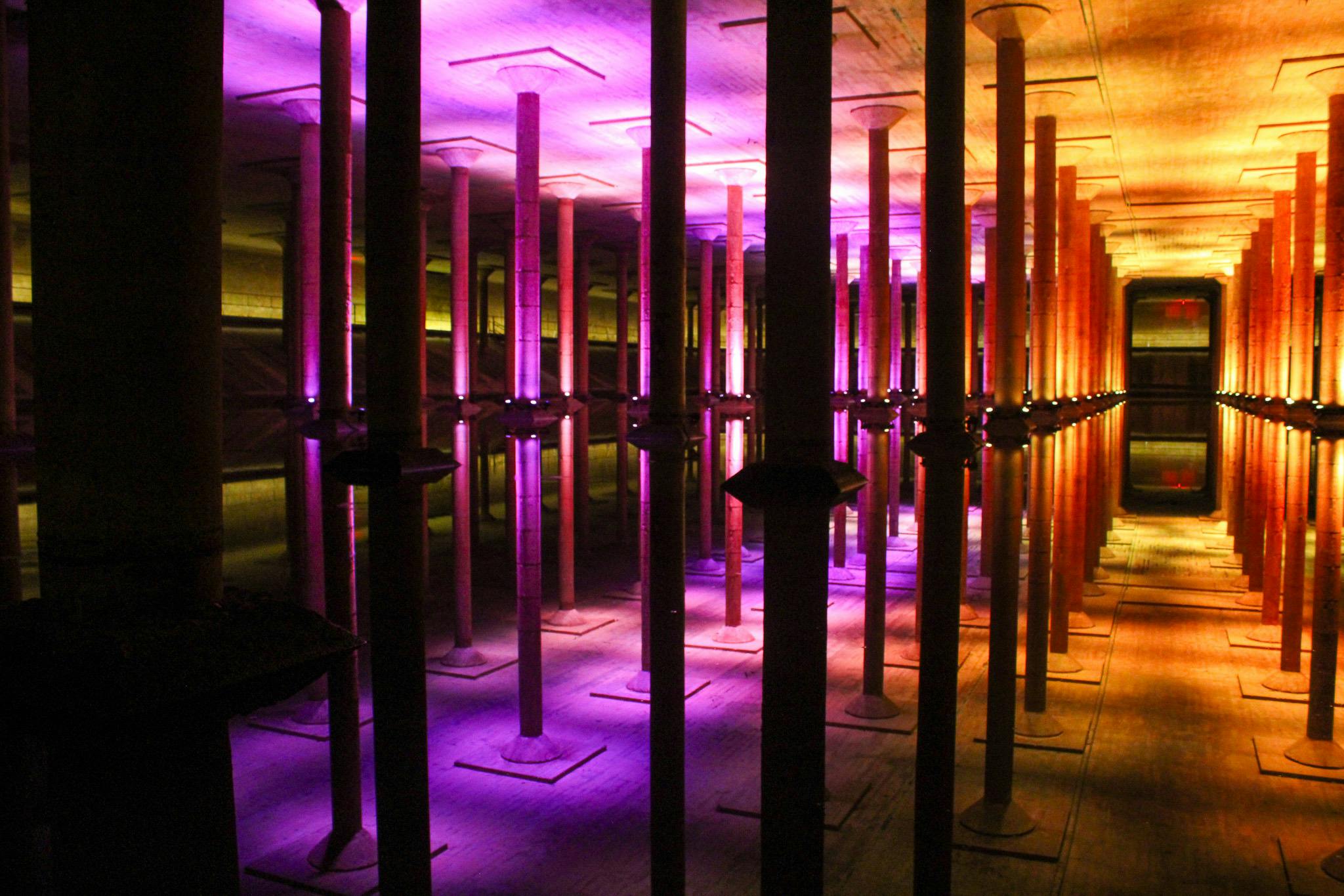 보라색과 주황색 조명으로 빛나는 높은 기둥이 있는 커다란 지하 홀이 아래 물에 반사됩니다.