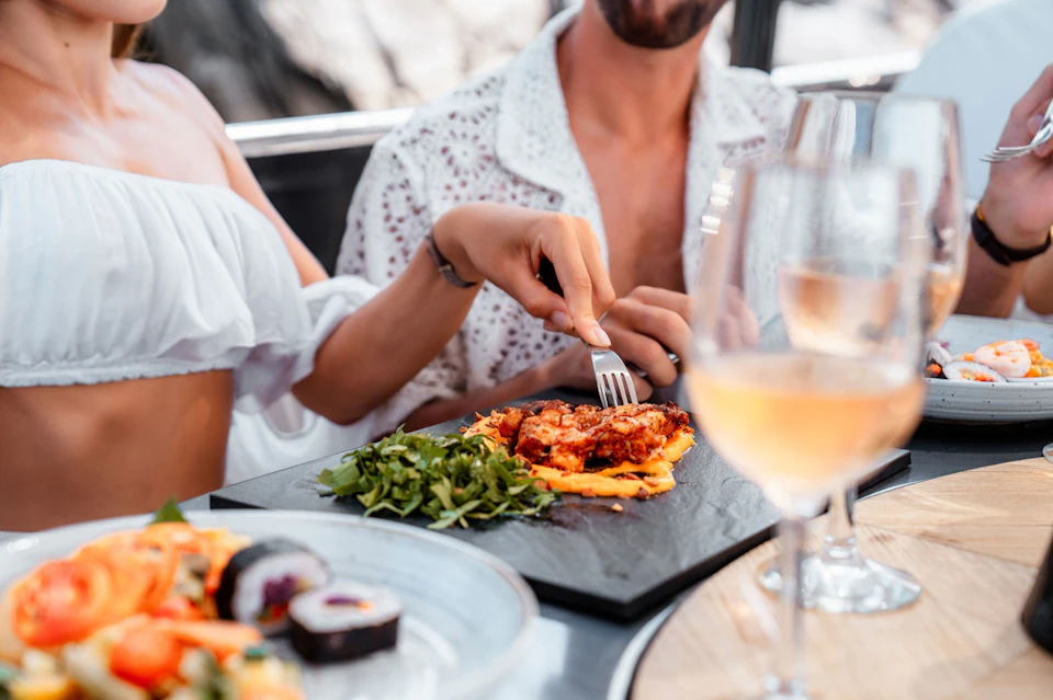 Two people are dining, with a hand using a fork to eat a dish beside a plate of sushi, and glasses of rosé wine visible.