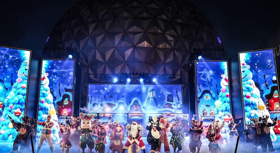 A group of performers in various festive costumes on a stage with a large geodesic dome and winter-themed decorations.