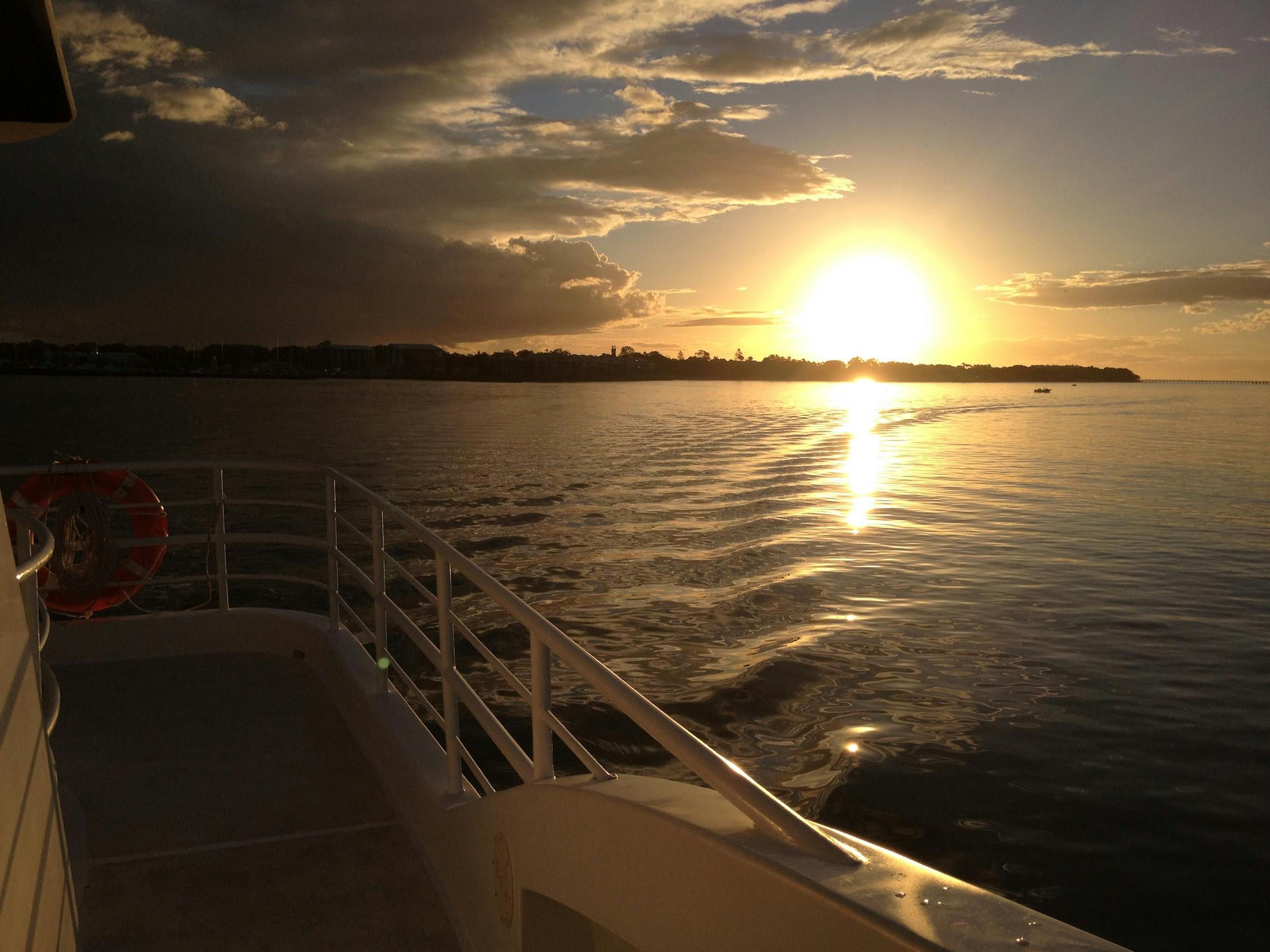 Sunset over calm water viewed from a boat, with the shoreline and buildings visible in the distance under a cloudy sky.