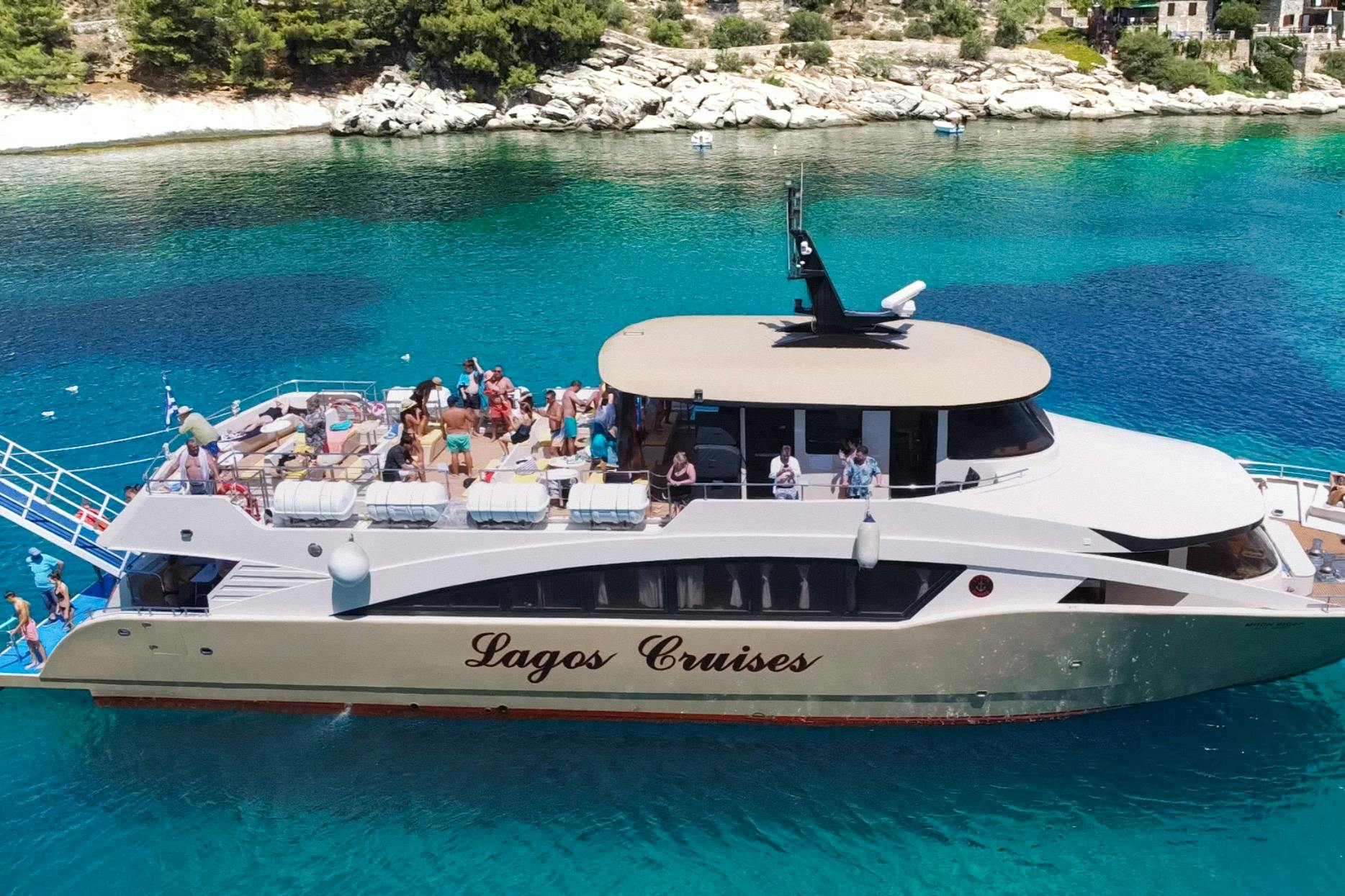 Un vaixell de creuer amb "Lagos Cruises" escrit al costat, que transporta persones, està ancorat en aigües blaves clares prop d'una costa rocosa.