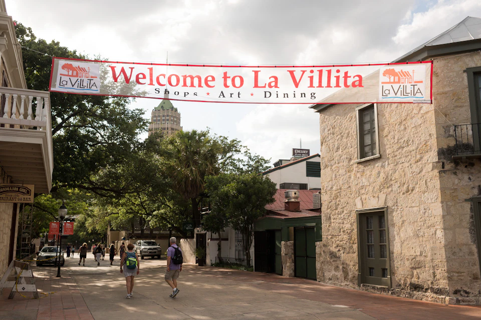 Street scene in La Villita with a red and white banner that reads "Welcome to La Villita, Shops, Art, Dining." People are walking.