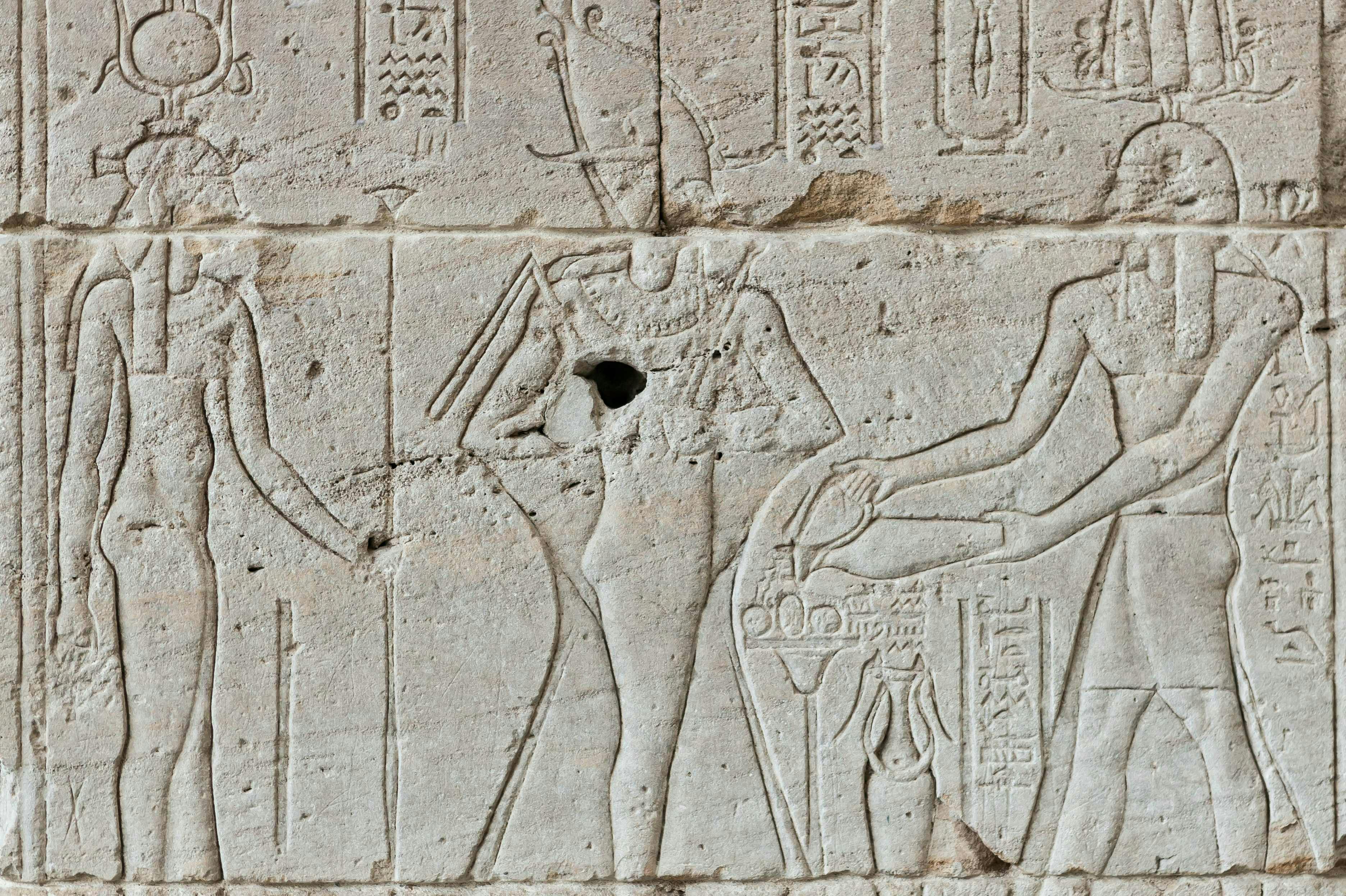 Staroegyptská nástěnná řezba zobrazující profil božstva obklopeného hieroglyfy a držící obřadní předměty.
