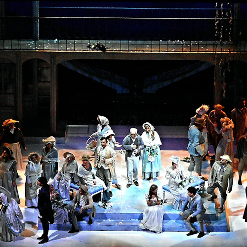 En teatralisk scen med många skådespelare i tidstypiska kostymer, stående och sittande mitt i en detaljerad uppsättning på flera nivåer.