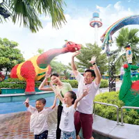 Eine lächelnde vierköpfige Familie hebt freudig die Hände neben bunten Lego-Skulpturen, darunter ein roter und grüner Dinosaurier, in einem Park.