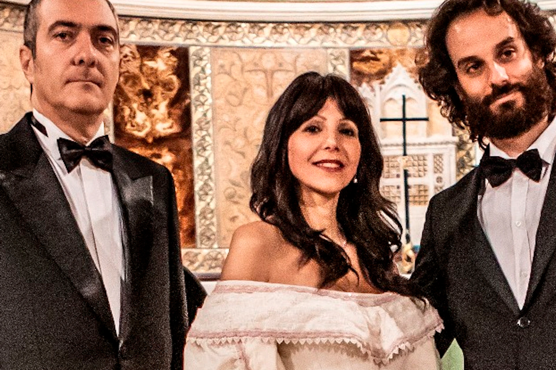 A group of musicians and singers in formal attire perform in an ornate church with frescoed walls and arched architecture.