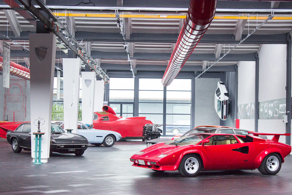 Several luxury sports cars, including a red Lamborghini, in a spacious, well-lit garage with large windows and a red ventilation pipe.