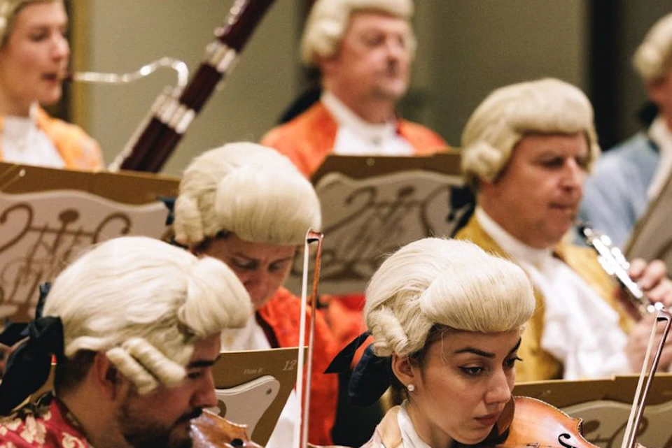 Orchestra musicians in period costumes with wigs, playing string and wind instruments. Music stands have ornate sheet music.
