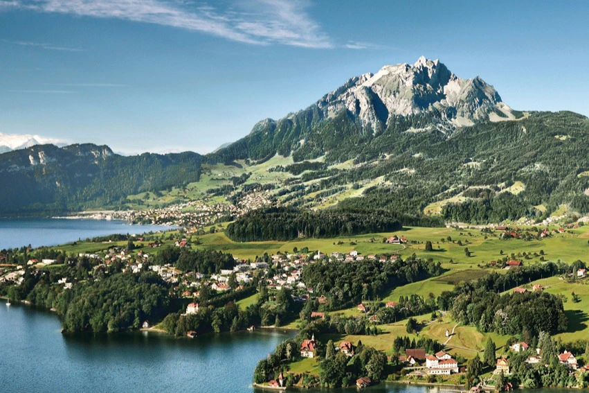 Scenic landscape of a lakeside village with green fields, scattered houses, and a large mountain range in the background under a blue sky.