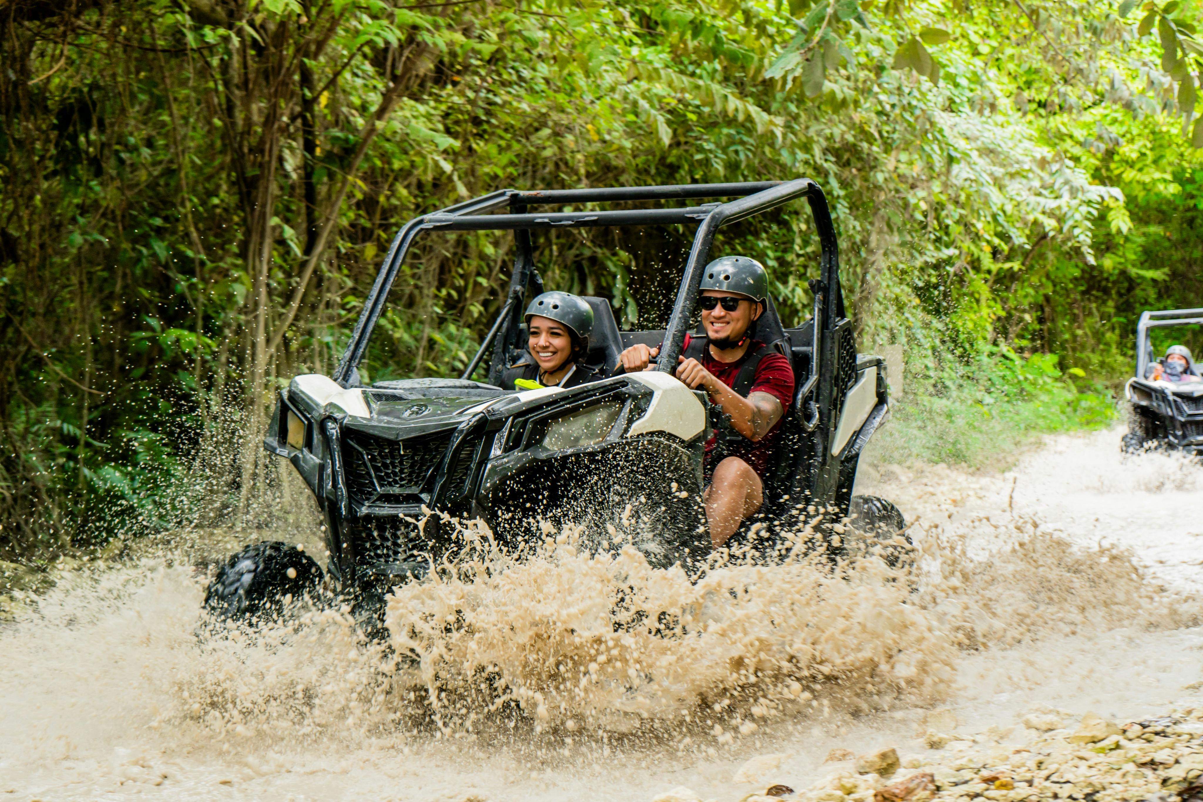 Two people in a utility terrain vehicle drive through a muddy puddle in a forest setting, wearing helmets and smiling.