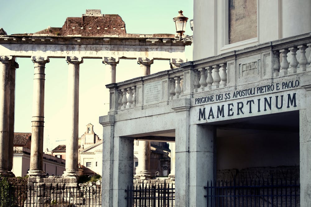 Ancient Roman columns and ruins behind Mamertine Prison with a sign in Italian, iron fence in the foreground, under a clear sky.