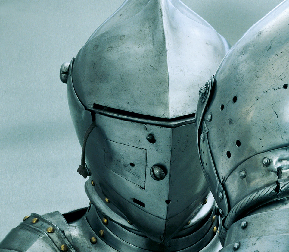 Close-up of two suits of medieval knight armor, featuring metal helmets with narrow eye slits and riveted joints.