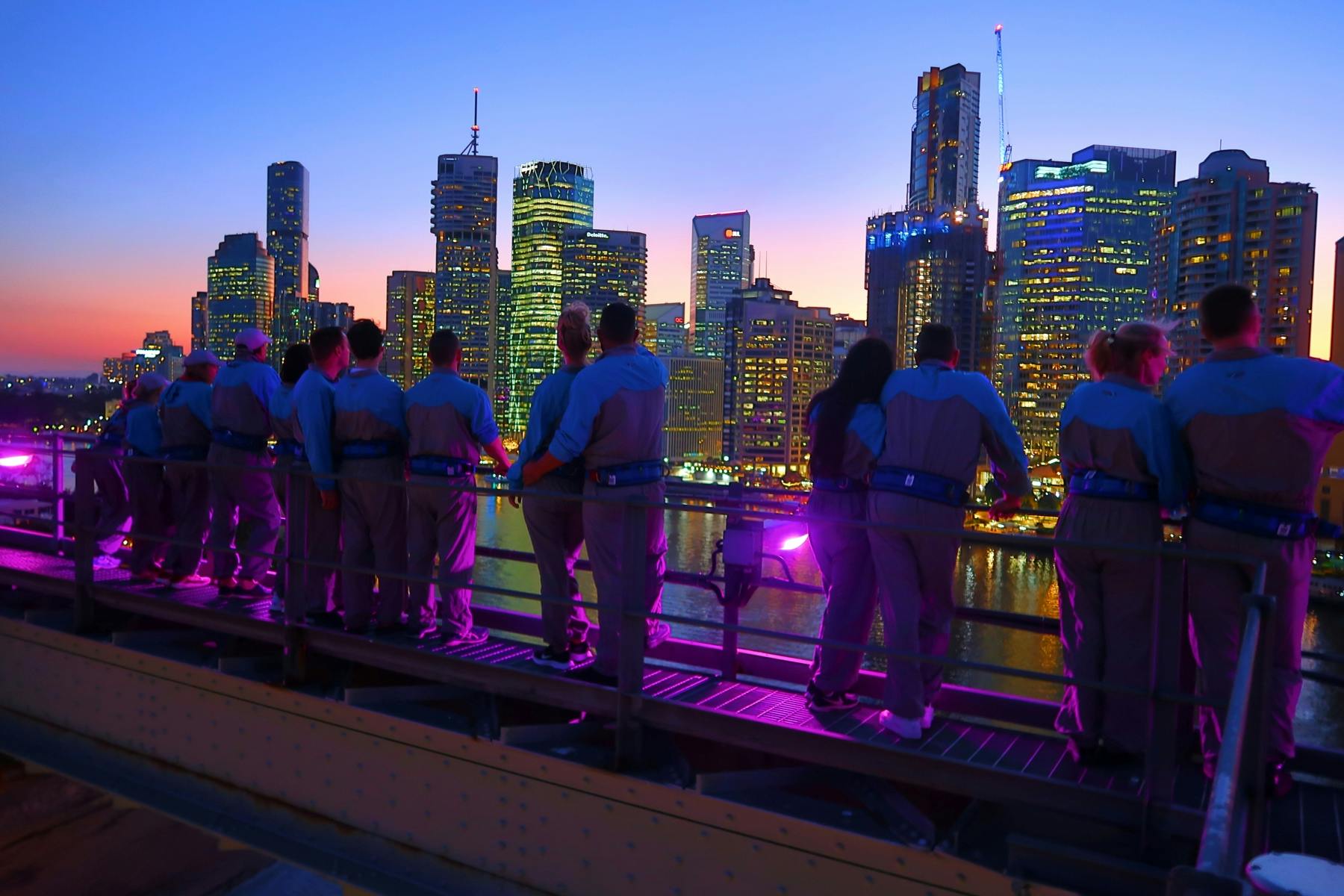 People in harnesses overlooking a city skyline at twilight from an elevated platform. Buildings are illuminated against the evening sky.