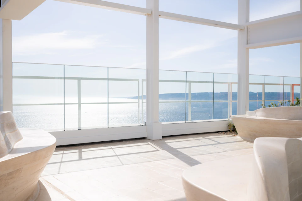 A spacious, modern terrace with glass railings overlooks a serene body of water and distant coastline under a clear sky.