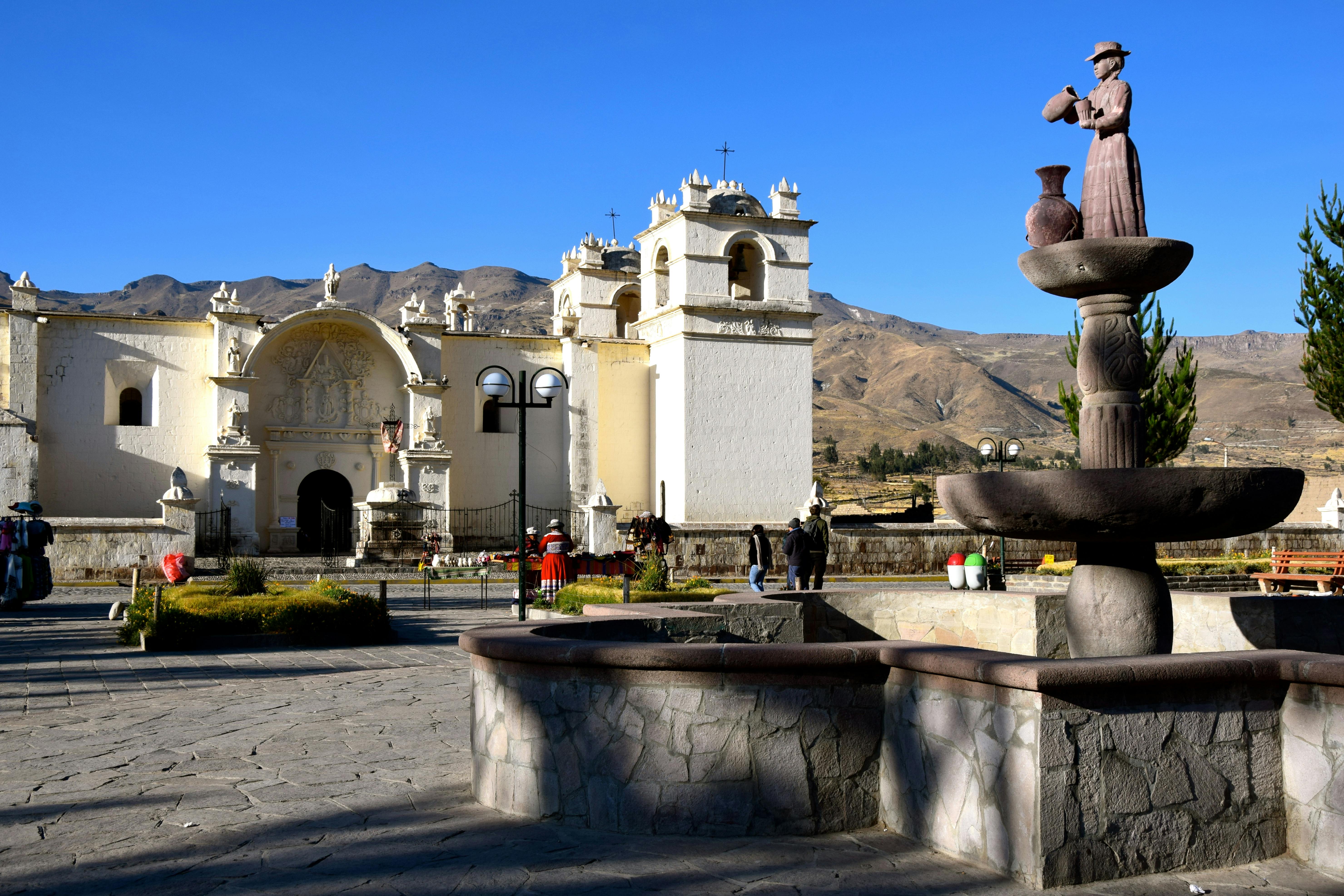 Белая церковь с колокольнями на площади, горы на заднем плане, гуляющие люди, каменный фонтан на переднем плане.