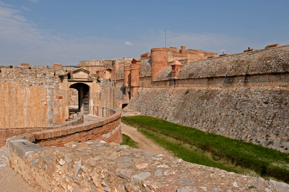 Historic fortress with stone walls, a guard tower, grassy moat, and an entrance gate under a clear blue sky.