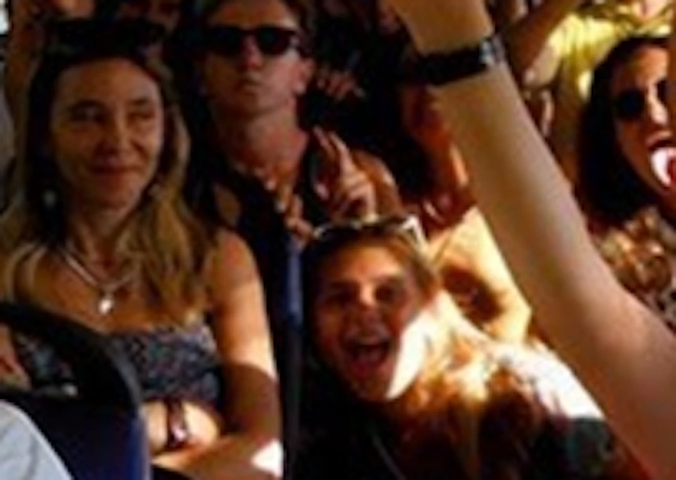 A group of people of various ages, smiling and raising their hands, sits inside a yellow tour bus with open windows.
