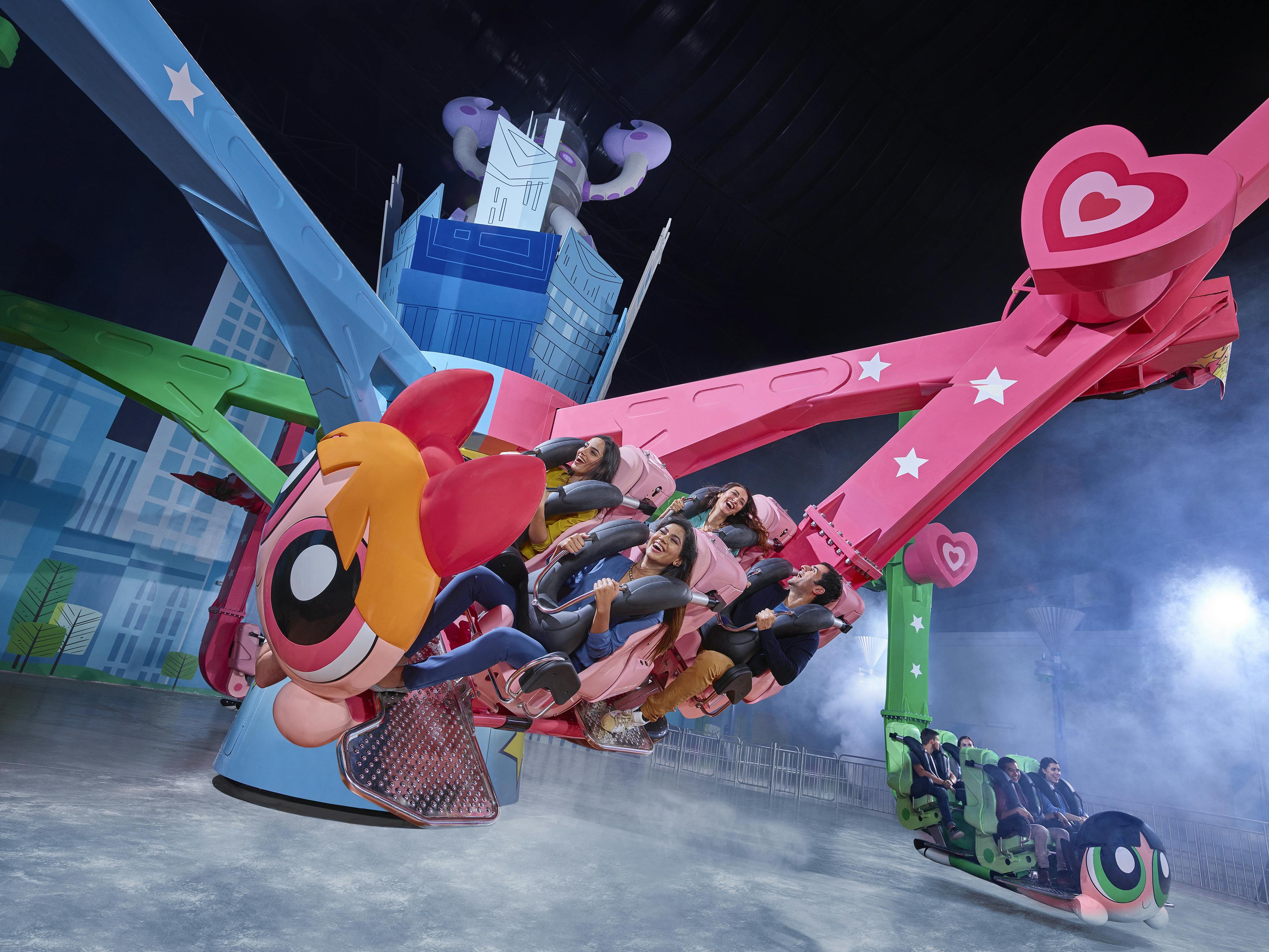 Άνθρωποι που απολαμβάνουν μια πολύχρωμη βόλτα σε πάρκο ψυχαγωγίας με θέμα γύρω από τα Powerpuff Girls, με ζωντανά μπλε και ροζ χέρια και καρδιές.