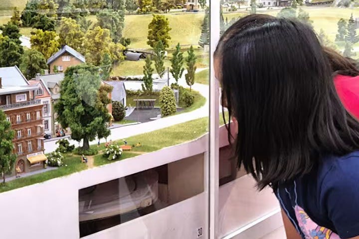 A person with black hair looks closely at a glass display of a miniature countryside scene with houses, trees, and roads.