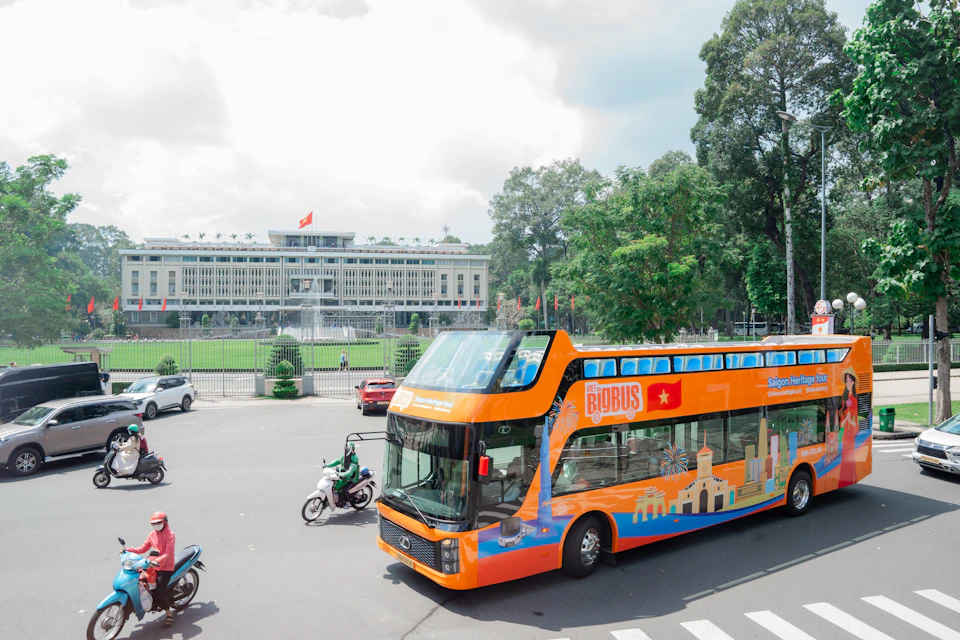 A bright orange double-decker bus in a street near a large building with a fountain and green trees in the background.