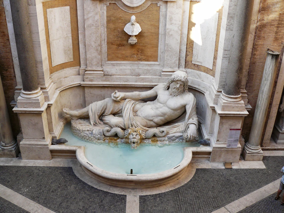 A marble statue of a reclining man in a small fountain with architectural details and a bust above the statue.