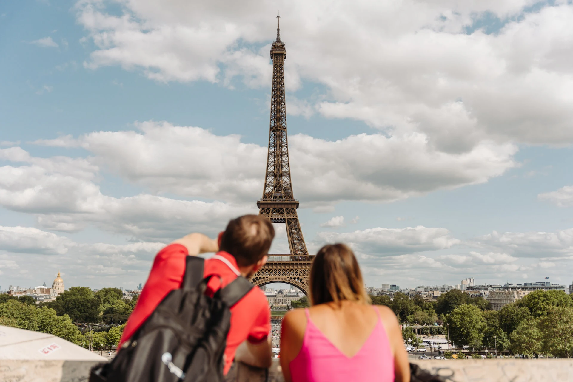 Two people sitting with their backs to the camera, looking at the Eiffel Tower on a sunny day with scattered clouds.