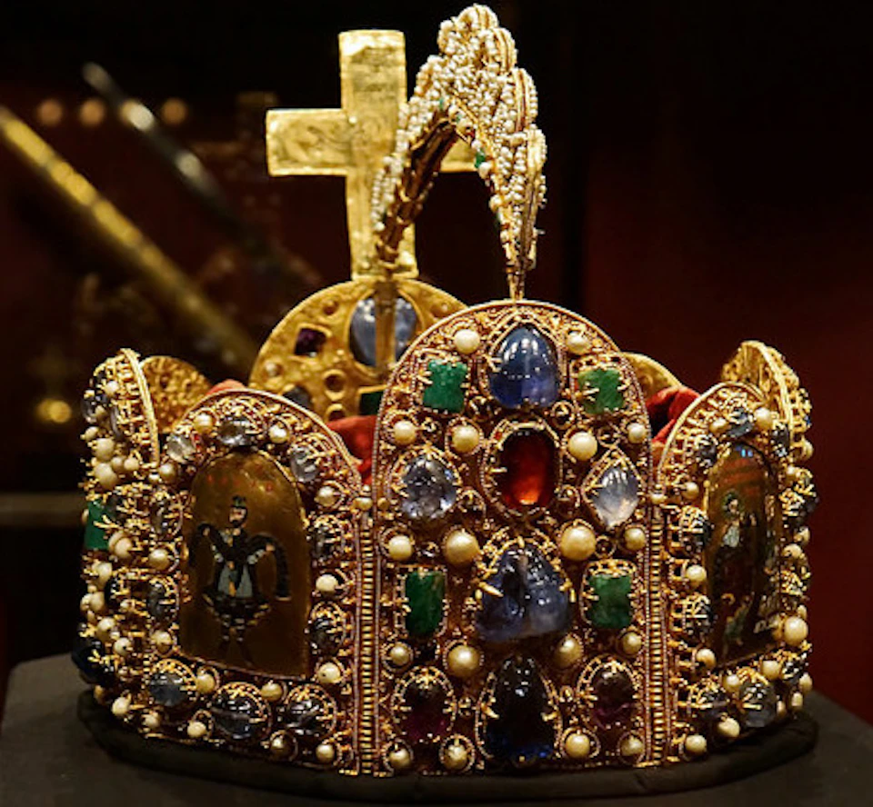 An ornate gold crown adorned with gems, pearls, and religious icons, topped with a cross.