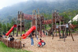 Children and adults at a wooden playground with slides and climbing structures, surrounded by grass and mountains.