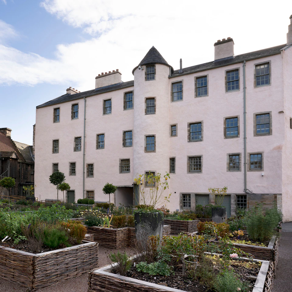 A historical stone building with multiple small windows, surrounded by a garden with various raised plant beds.