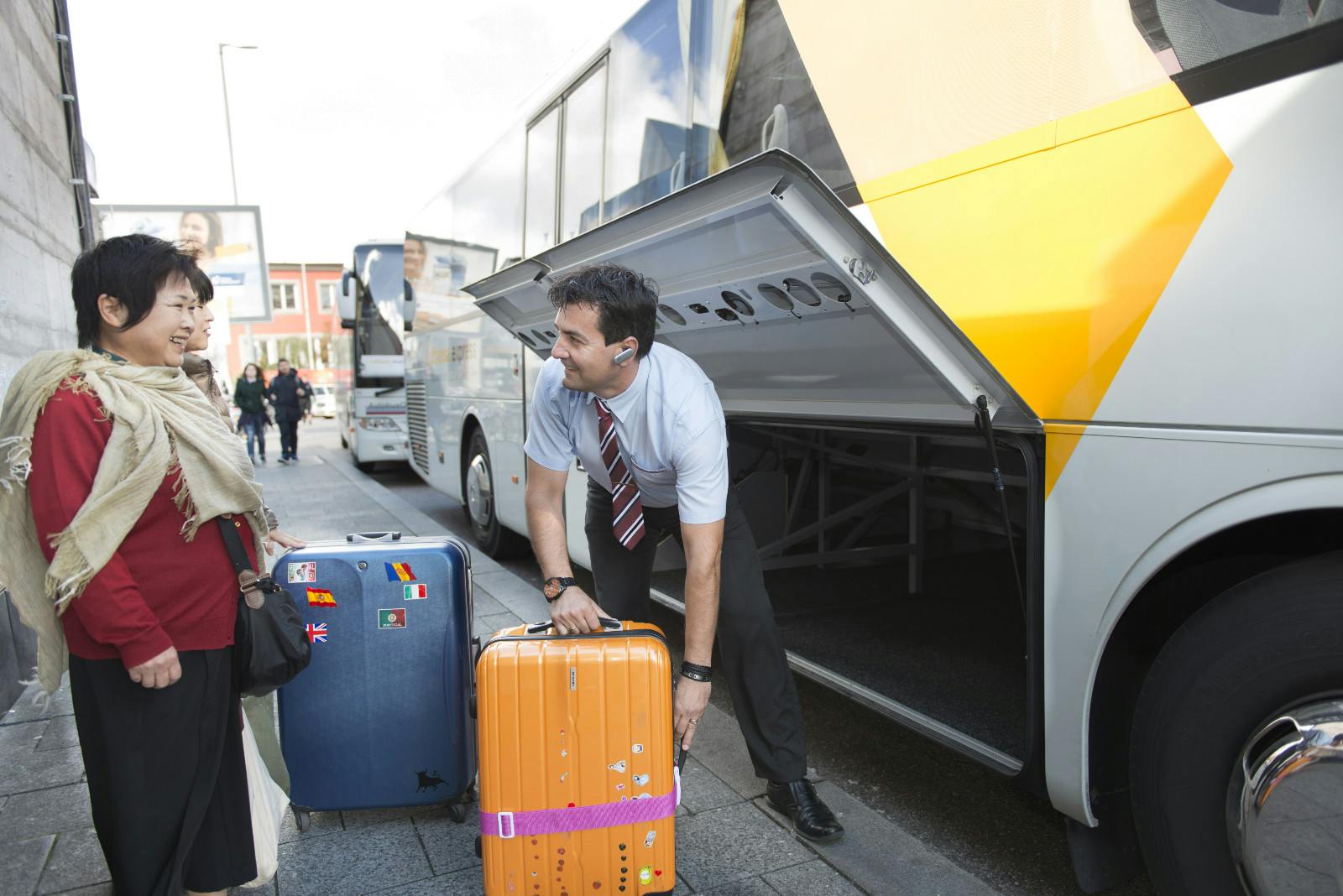 A man in a uniform loads suitcases onto a bus while talking to a smiling woman with a red top and beige shawl.