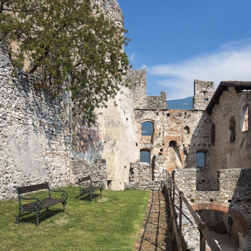 Stone ruins with arches and windows, grassy area with benches, tree, iron fence, and a clear blue sky.