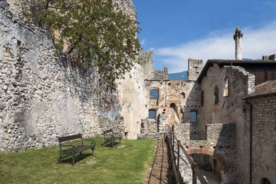 Stone ruins with arches and windows, grassy area with benches, tree, iron fence, and a clear blue sky.
