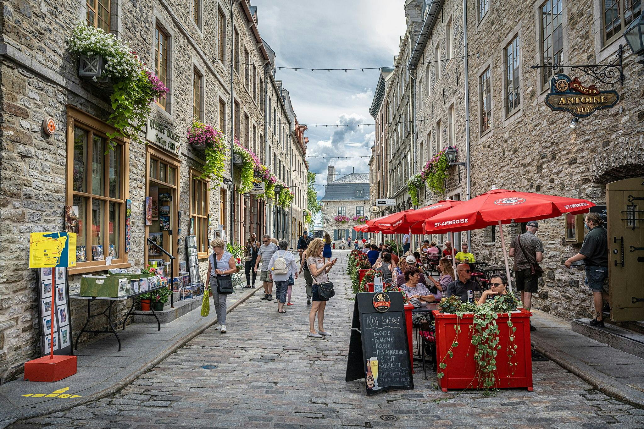 Eine Straße mit Kopfsteinpflaster, gesäumt von Steingebäuden, kleinen Geschäften, roten Café-Schirmen, Blumen und Menschen, die spazieren gehen und essen.
