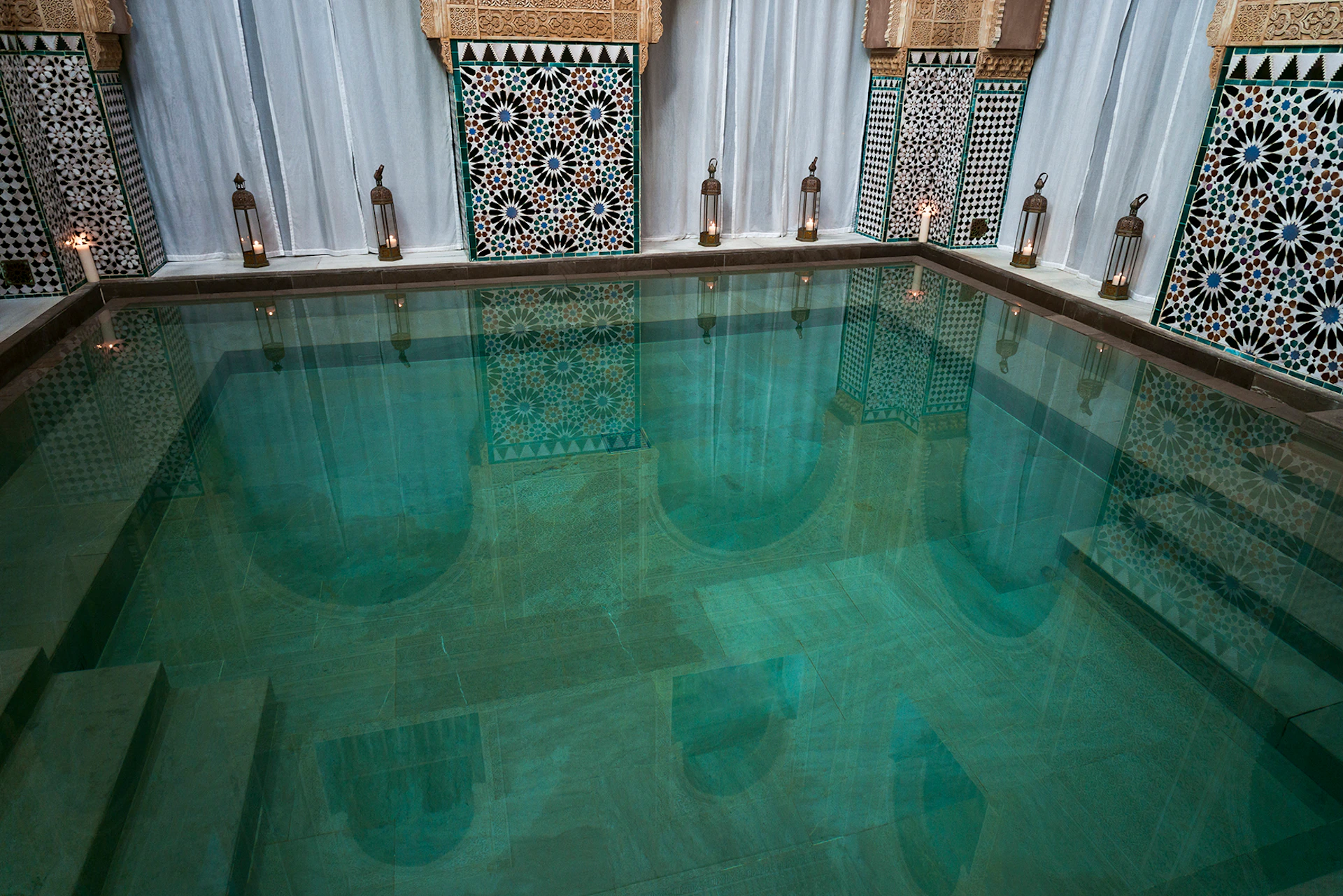 A serene indoor pool with clear water, surrounded by ornate tiles and lanterns, framed by white curtains.