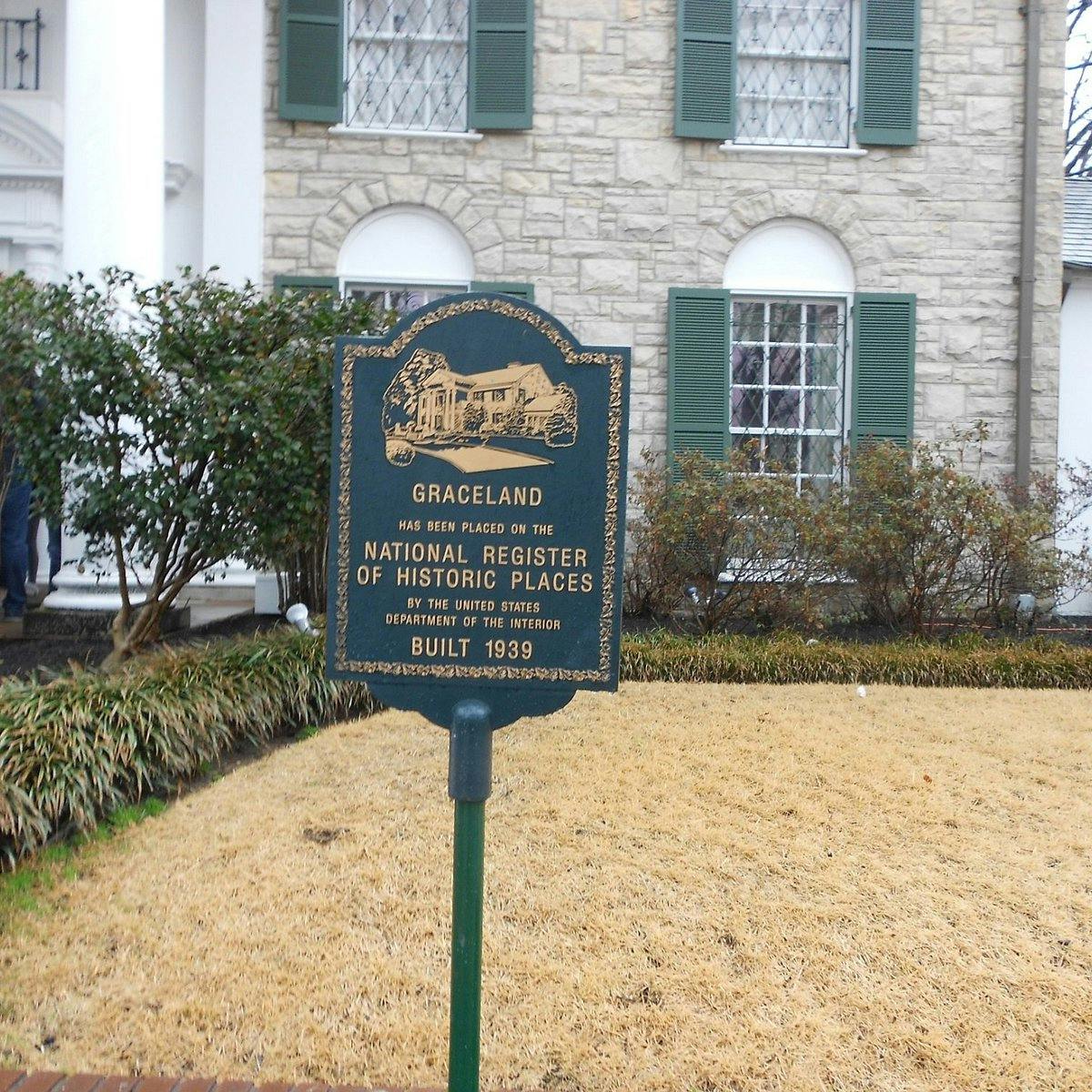 A plaque in front of a house with stone walls, green shutters, and a manicured lawn, indicating Graceland's historic status.