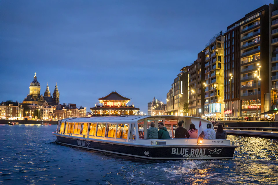 A canal boat with passengers navigates a waterway in a city during the evening, surrounded by illuminated buildings and landmarks.