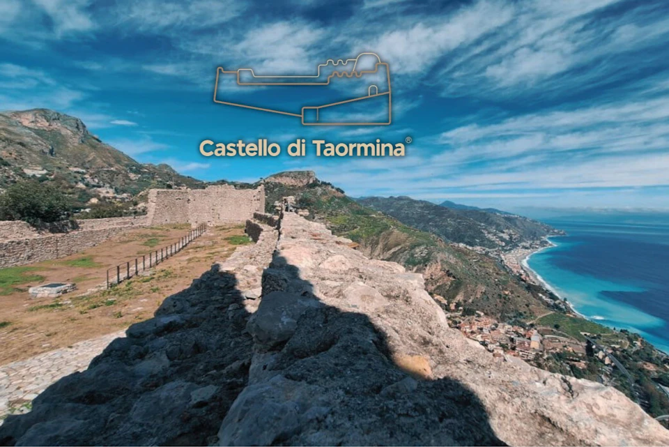 View of Castello di Taormina ruins with a coastal landscape, blue sky, and a shadow on the stone path in the foreground.