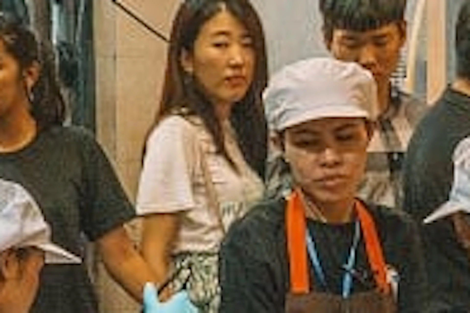 Busy street food stand with chefs in black shirts and white hats preparing meals, while customers wait in line. Signs in Thai above.