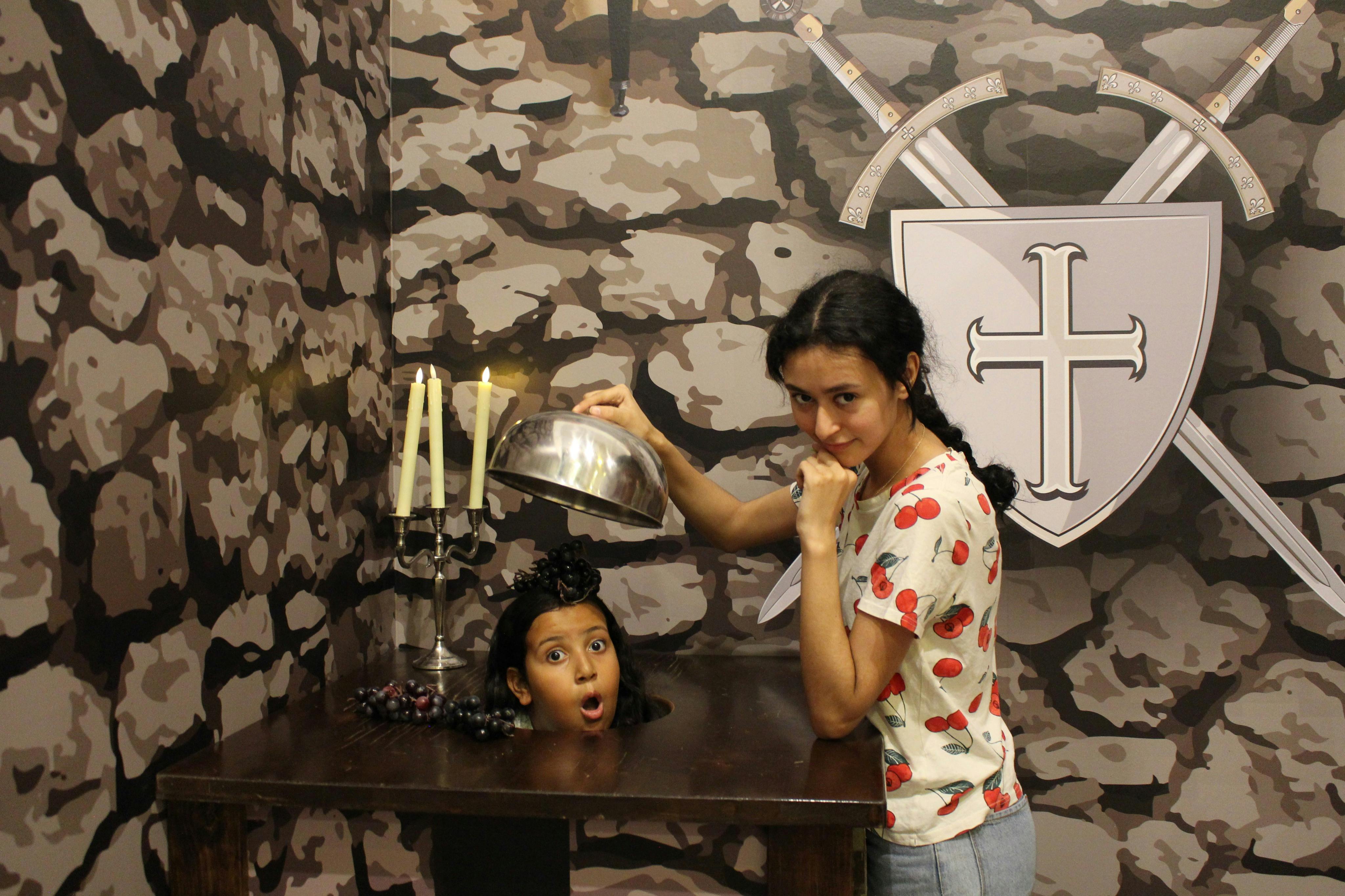 Una dona amb una samarreta amb estampat de cireres sosté una cúpula platejada sobre una taula amb el cap d'un nen que apareix a través de la superfície, semblant sorprès. El fons té un disseny de camuflatge i un escut amb l'emblema de les espases.