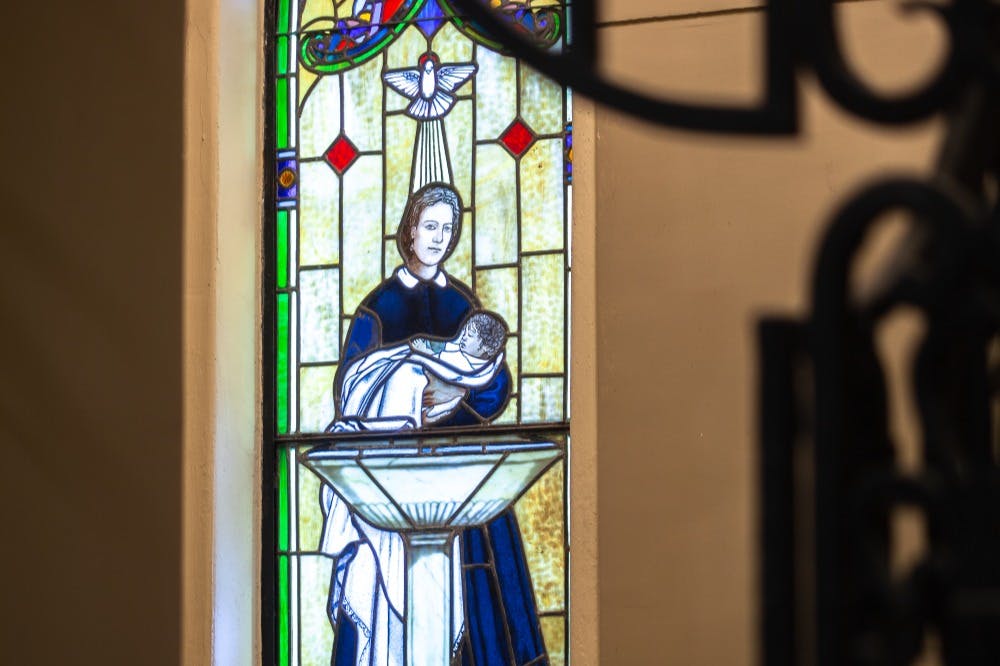 Βιτρό που απεικονίζει μια φιγούρα που κρατά ένα βρέφος πάνω από μια γραμματοσειρά βάπτισης, με ένα περιστέρι από πάνω τους.