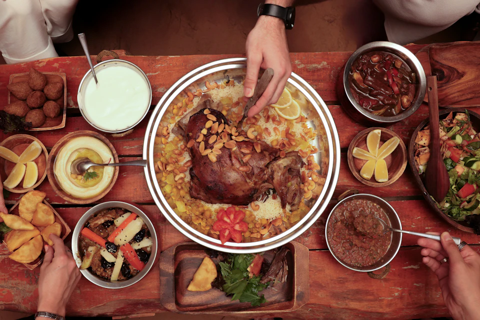 A hand serving a roast meat dish with almonds and vegetables surrounded by various side dishes on a wooden table.