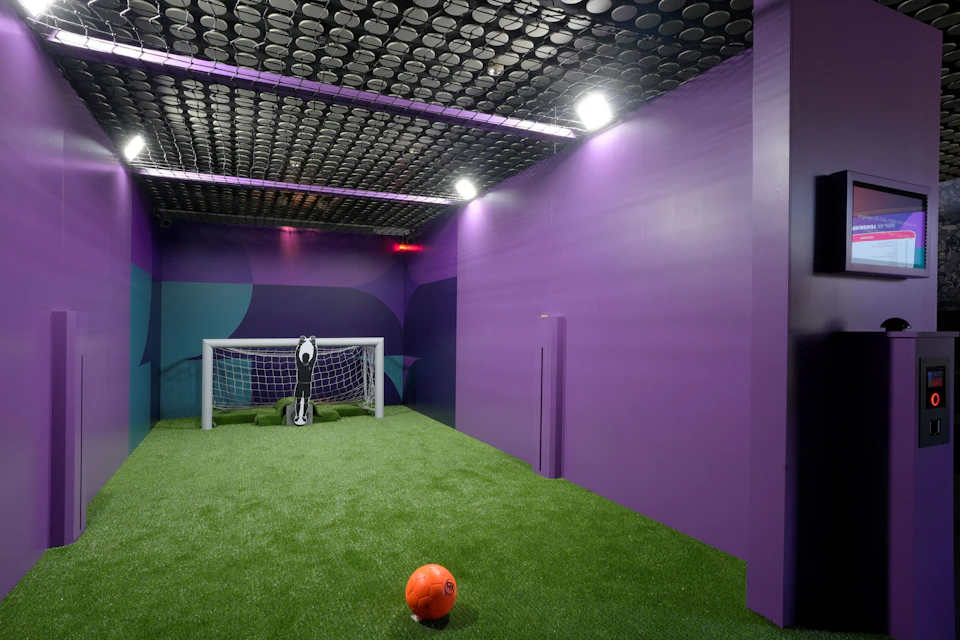 A small indoor soccer field with artificial turf, purple walls, a goal net, and an orange soccer ball on the ground.