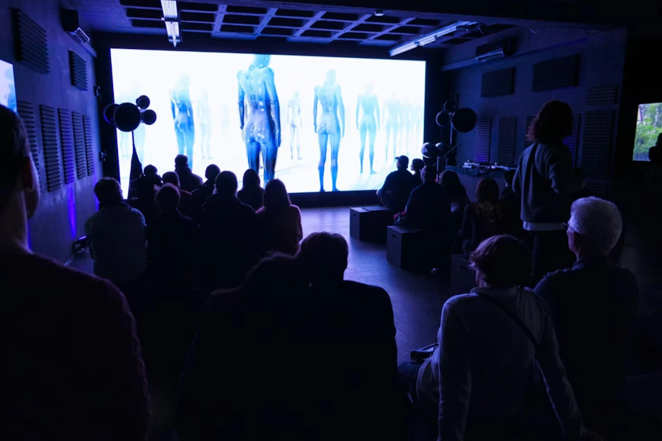 A group of people seated in a dimly lit room facing a large screen showing abstract humanoid figures in blue tones.